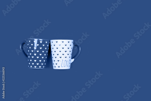 Two cups painted with hearts and the word Love written on one of them on deep blue background with copy space for text. Color trend of 2020 concept.