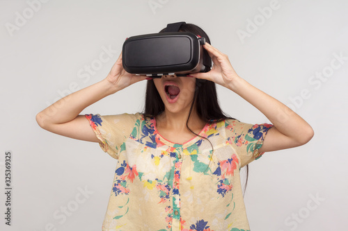 Surprised woman wearing VR glasses and watching virtual world with amazed shocked expression, playing video game, experiencing strong emotions while using simulator. indoor studio shot gray background