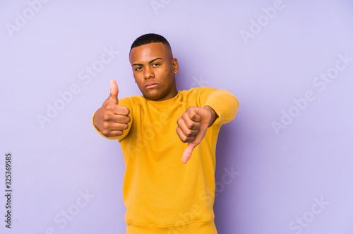 Young latin man isolated on purple background showing thumbs up and thumbs down, difficult choose concept
