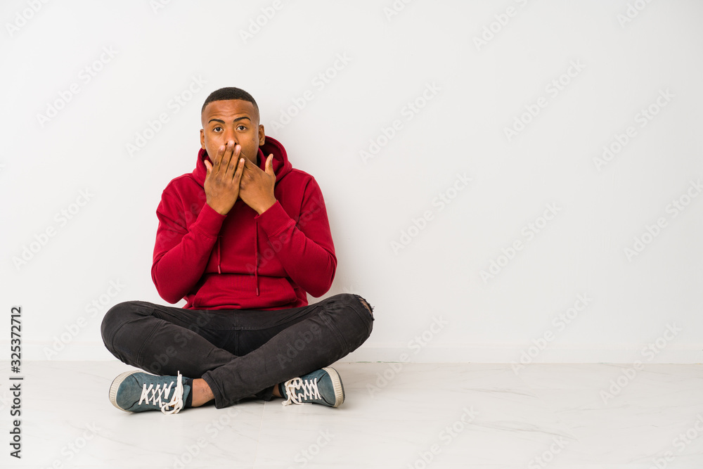 Young latin man sitting on the floor isolated shocked covering mouth with hands. Young latin man sitting on the floor isolated shocked covering mouth with hands.