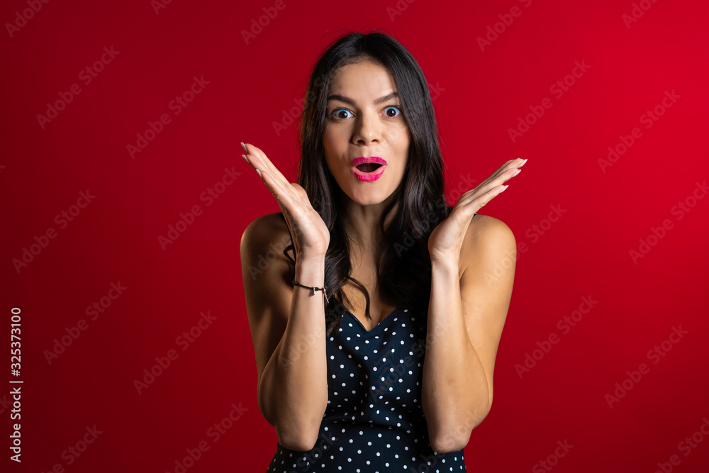 Obraz premium Pretty female shocked model on red background. OMG, wow effect. Hispanic girl with long hair with delight. Surprised excited happy woman in dress.