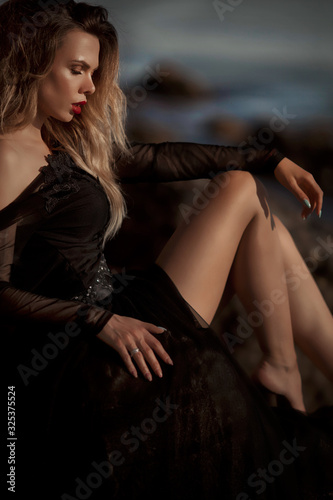 Young woman in long transparent black dress among beach and sea. Wind blows hem of her dress.