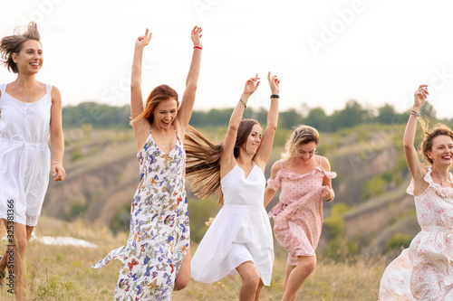 The company of cheerful female friends have a great time together on a picnic in a picturesque place overlooking the green hills. Girls in white dresses dancing in the field