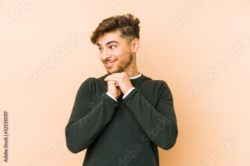 Young arabian man isolated on a beige background keeps hands under chin, is looking happily aside.