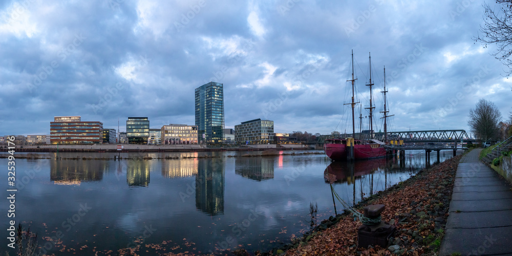 Naklejka premium panorama of the Überseestadt in Bremen, Germany with office building, sail boat and perfect reflection on the river weser during blue hour