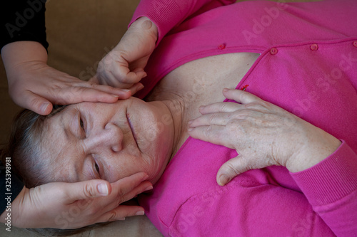 Seriously ill elderly woman in pink blouse is lying on sofa. Girl nurse in white medical gown from center of social assistance and protection comforts dying woman and strokes her head with hands