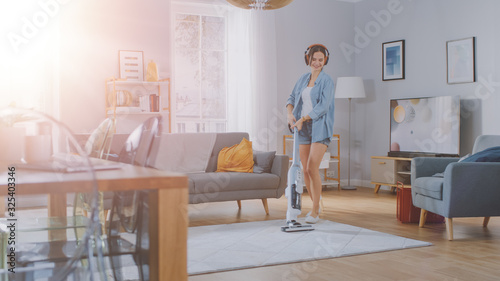 Shot With Sun Flare of a Young  Woman is Listening to Music on Her Headpones, Dancing and Vacuum Cleaning a Carpet in a Cozy Room at Home. She Uses a Cordless Vacuum. She's Happy and Joyful. © Gorodenkoff