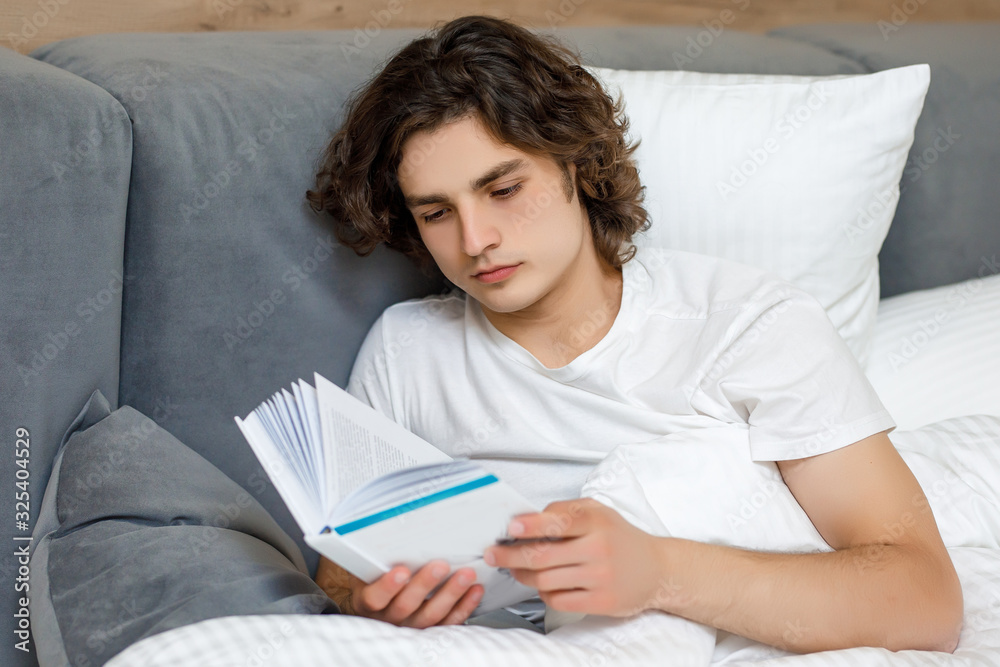 Handsome young man laying on his bed reading a book, resting at his bedroom Handsome young man laying on his bed reading a book, resting at his bedroom