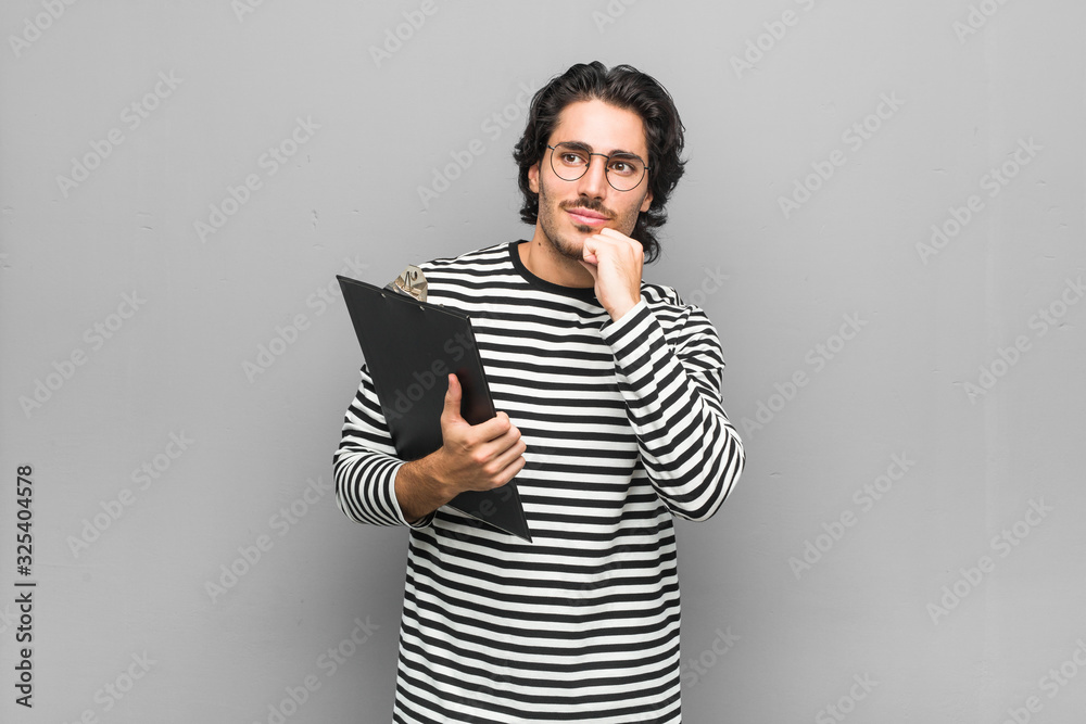 Young employee man holding an inventory keeps hands under chin, is looking happily aside. Young employee man holding an inventory keeps hands under chin, is looking happily aside.