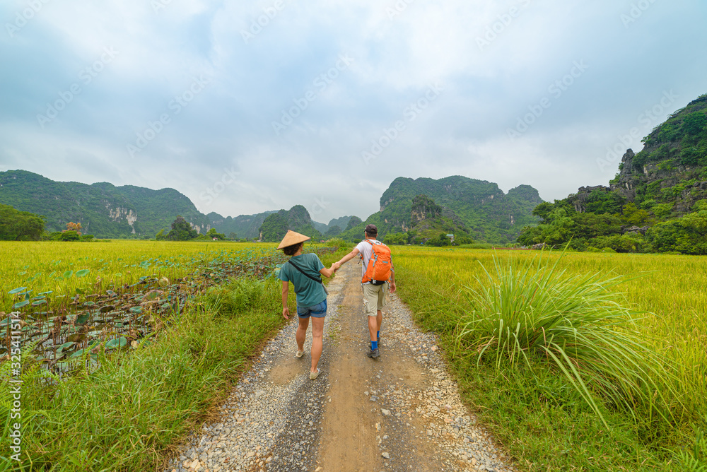 Couple walking hand on countryside road among rice fields. Man with backpack and Woman with vietnamese hat having fun traveling together on vacation. Tam Coc Trang An Ninh Binh karst mountain range.