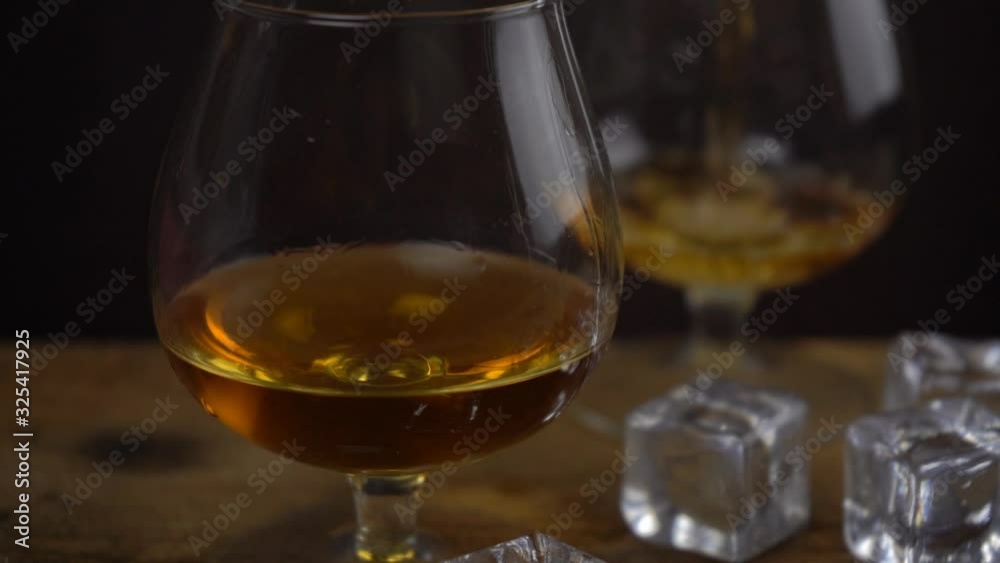 Cognac \ whiskey pouring into glass, golden color strong spirit alcohol in cognac glasses on dark black background, slow motion footage Cognac \ whiskey pouring into glass, golden color strong spirit alcohol in cognac glasses on dark black background, slow motion footage