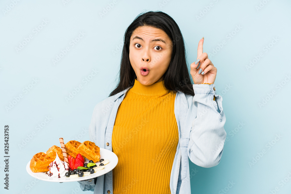 Young chinese woman holding waffle isolated on blue background laughs out loudly keeping hand on chest. Young chinese woman holding waffle isolated on blue background laughs out loudly keeping hand on chest.