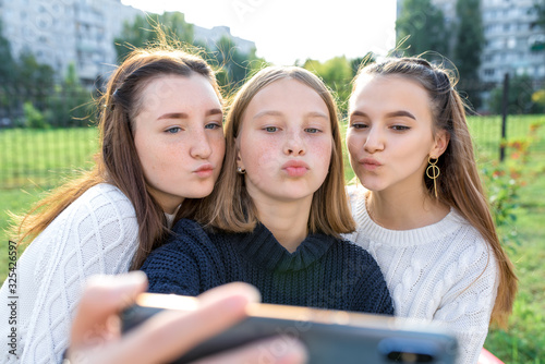 Three girls schoolgirls teenagers summer city. Photography on phone, selfie photo, online app smartphone, social networks on Internet. Sweater casual wear, air kiss, emotions relaxation fun pleasure.