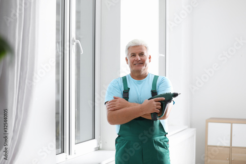 Mature repairman with electric screwdriver near plastic window indoors