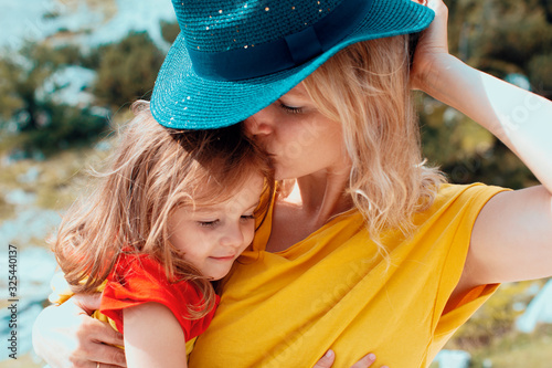 A young mother showing love hugging and kissing her daughter outdoors in the sun, covering her with hat