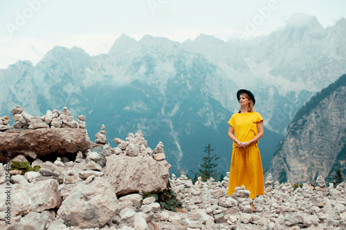 Portrait of a young woman in yellow standing in front of a  big mountain ridge with stone sculptures and holding a stone photo