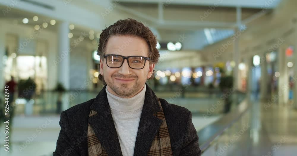 Portrait of Caucasian attractive man in coat and glasses standing at shopping mall passage, turning face and smiling joyfully to camera. Close up of male customer looking happy at big shop corridor. Portrait of Caucasian attractive man in coat and glasses standing at shopping mall passage, turning face and smiling joyfully to camera. Close up of male customer looking happy at big shop corridor.