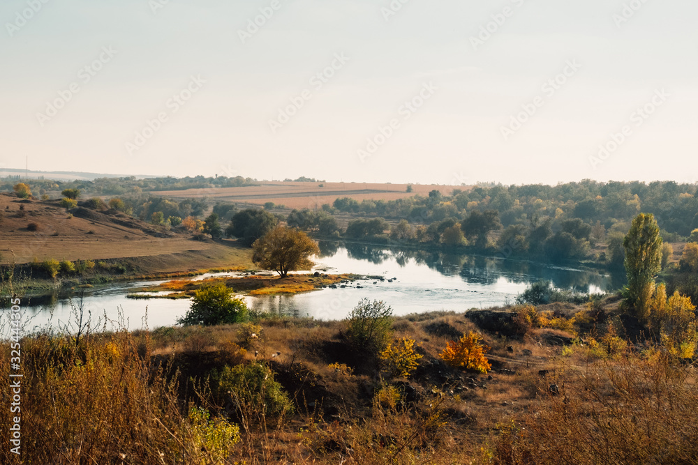 Meandering river, lush green rural forest and rocky stone cascade in warm sunlight. Colorful autumn landscape, Southern Bug, Ukraine. Beautiful fall nature with yellow trees. Mountain hills view.