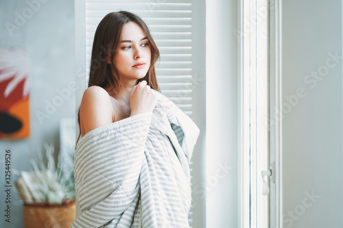 Young beautiful long brown-haired hair girl in blanket near window at bright modern interior morning routine