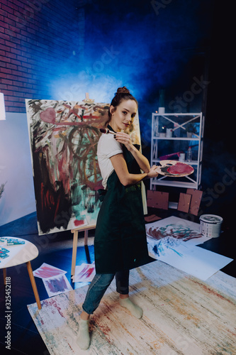A young female artist is creating an abstract painting. She is working in the loft space with dimmed lights. She is using oil paint and a brush.