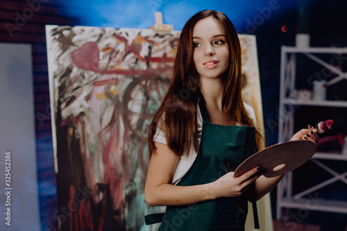 A young female artist is creating an abstract painting. She is working in the loft space with dimmed lights. She is using oil paint and a brush. © Owl-vision-studio