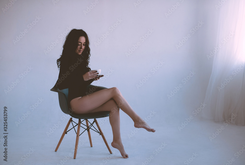 Fototapeta premium Beautiful woman drinking coffee sitting in chair wearing black elegant bodysuit in white room closeup. Good morning.