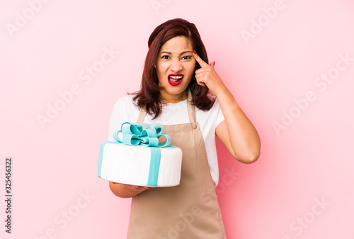Middle age latin woman holding a cake isolated on a pink background showing a disappointment gesture with forefinger.