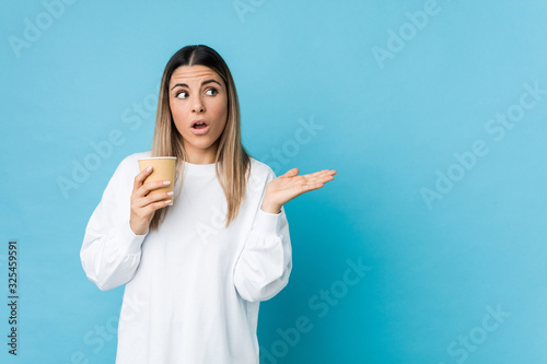 Young caucasian woman holding a takeaway coffee impressed holding copy space on palm.