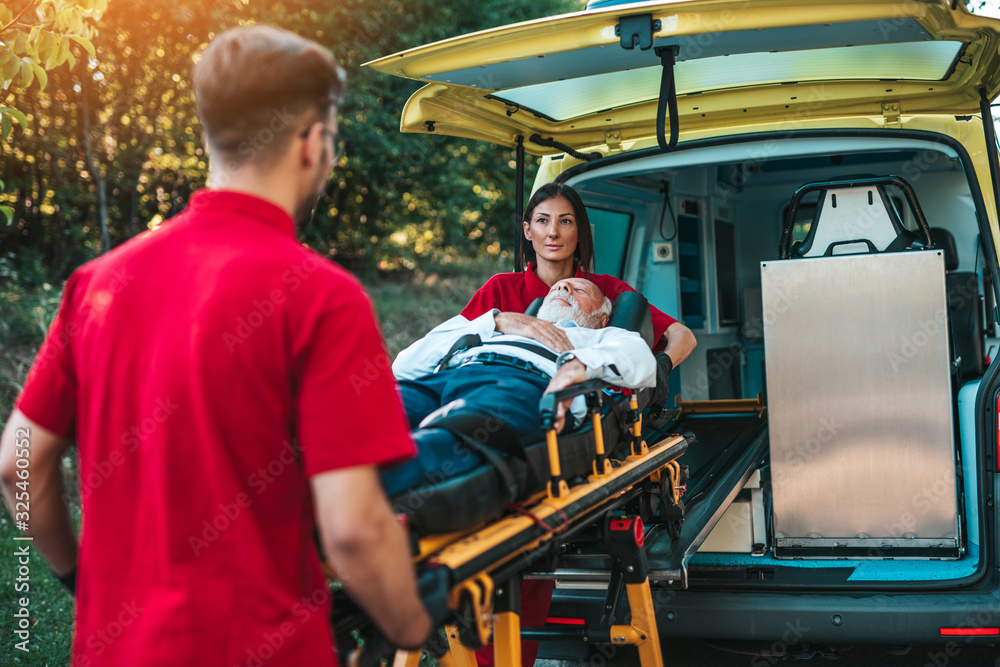 Emergency medical service at work. Paramedic is pulling stretcher with senior man with serious heart attack to the ambulance car. Help on the road. Drivers assistance concept. Emergency medical service at work. Paramedic is pulling stretcher with senior man with serious heart attack to the ambulance car. Help on the road. Drivers assistance concept.