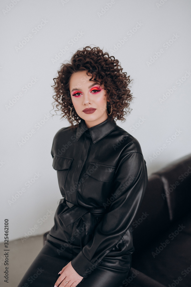 girl with bright makeup and curly hair in a black leather dress on a light background girl with bright makeup and curly hair in a black leather dress on a light background