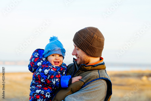 Happy father with his son on nature vacation in autumn, winter cold weather. Family spending time together. Dad holding his kid, little baby boy, carrying him outdoors, countryside on sunset.