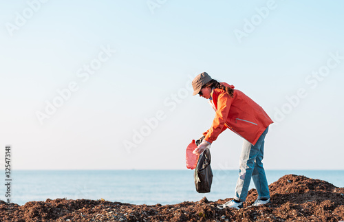 A young Caucasian woman in a jacket collects garbage in a plastic bag on the beach. In the background, the sea. Copy space