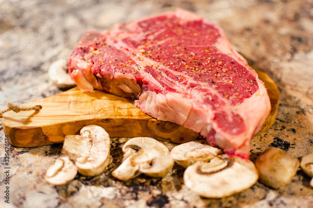 A T bone or Porterhouse raw steak on cutting board with mushrooms.  Blurred foreground.  Blurred background.  Neutral marbled surface.