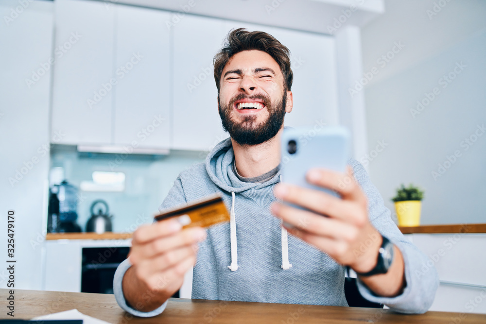 Portrait of cheerful young man laughing while paying online from home with credit card and smartphone