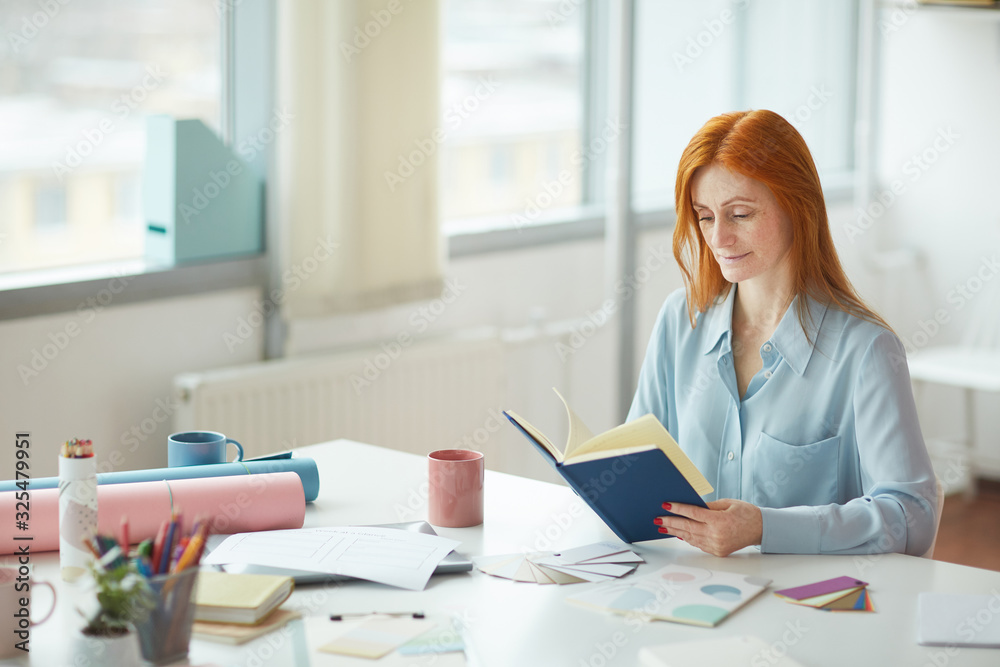 Portrait of freckled young woman reading journal while sitting at creative workplace by window, copy space Portrait of freckled young woman reading journal while sitting at creative workplace by window, copy space