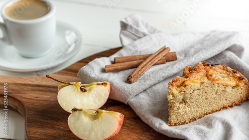 Apple pie slice lying on a grey cotton napkin. Wooden cutting board. There is cinnamon sticks  apple slices. And cup of coffee on the background. Wooden white background. 