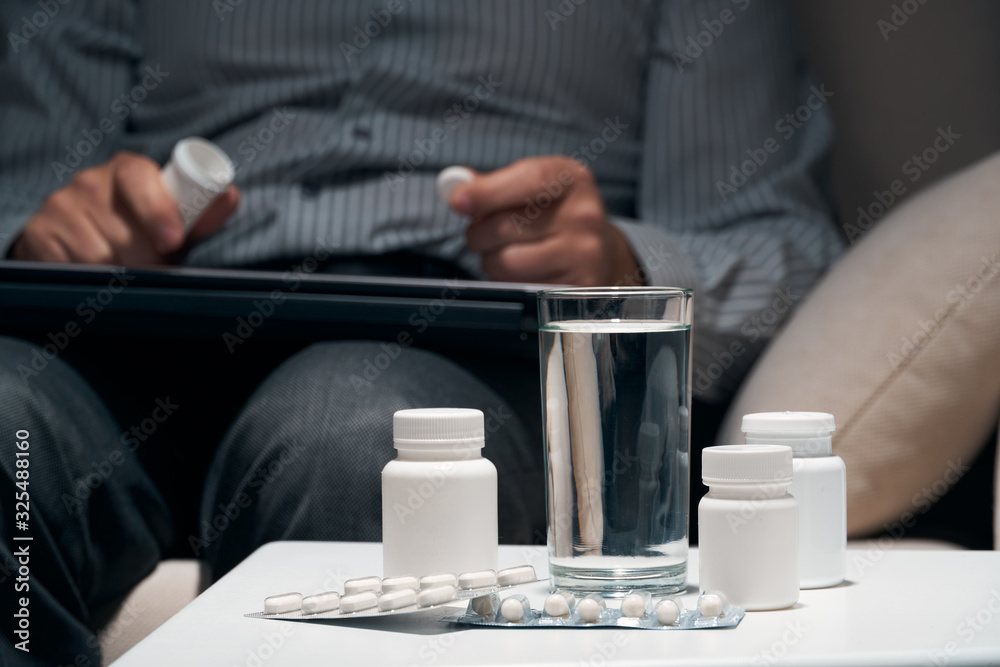 Overworked businessman sitting on the sofa and taking medication. Pills and glass of water near him.