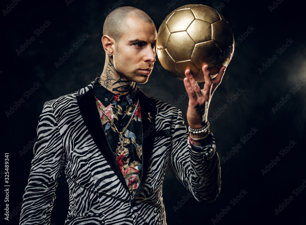 Plakat Trendy, vivid, tattooed, bald male model posing in a studio for the photoshoot wearing fashionable custom made zebra striped style tuxedo and rose patterned shirt, looking on a golden soccer ball