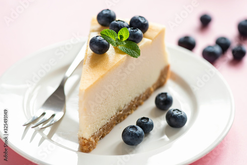 Classical Cheesecake Slice Served With Fresh Blueberries And Mint Leaf On White Plate, Pink Background. Tasty Sweet Dessert Food. Creamy Cake