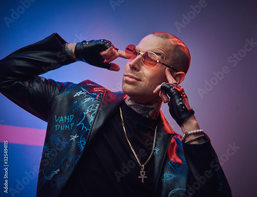 Creative, daring, tattooed, bald male rocker model posing in a studio for the photoshoot wearing fashionable designer black leather jacket, black shirt, red glasses, and jewelery, touching gead with photo