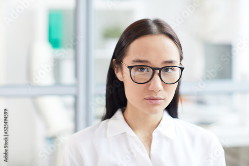 Head and shoulders portrait of young Asian businesswoman wearing glasses and looking at camera while posing in modern white office, copy space