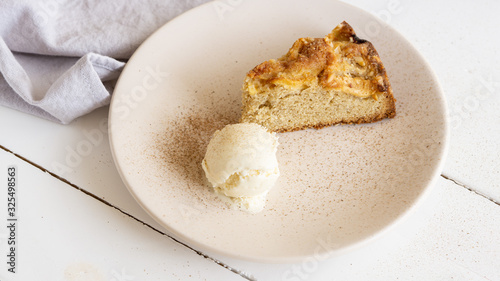 Apple pie slice with one bowl of vanilla ice cream on a round plate with some cinnamon in it. Near there is grey cotton napkin. White wooden table. 