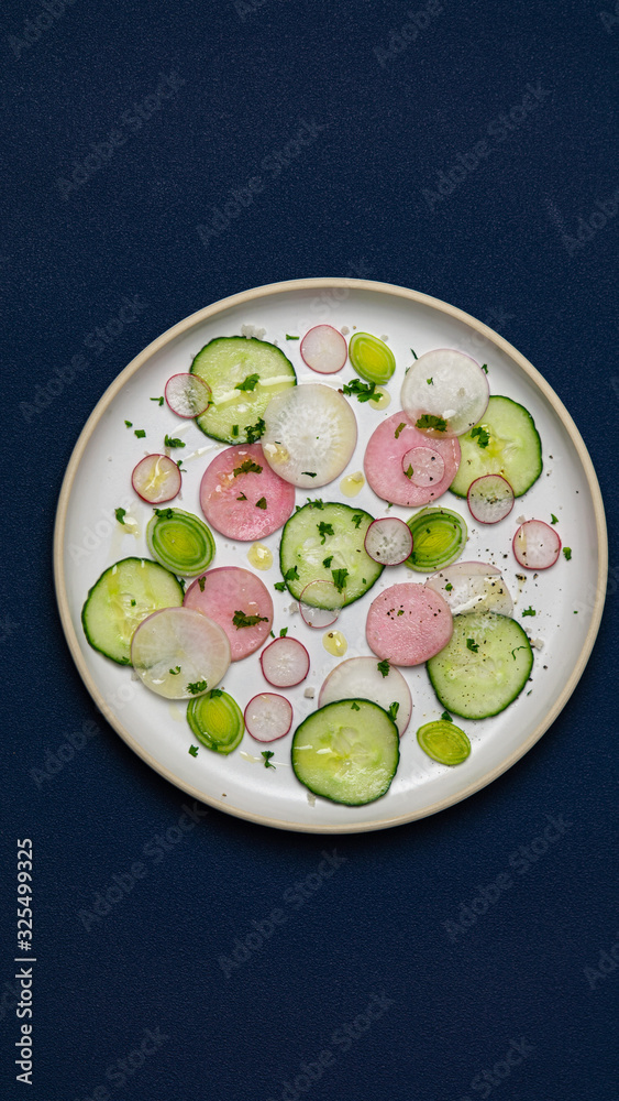 Cucumber and radish salad on a round plate on blue background. There are some persil and leek an a plate. 
