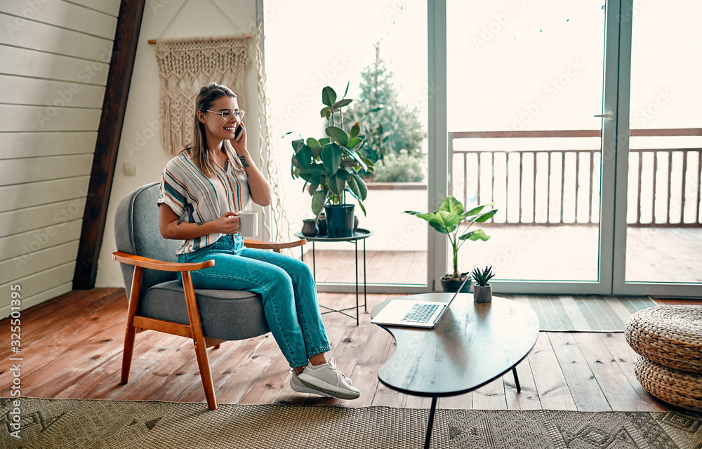 Fototapeta premium An attractive young woman in glasses is drinking coffee and talking on the phone while sitting in a comfortable chair at home.