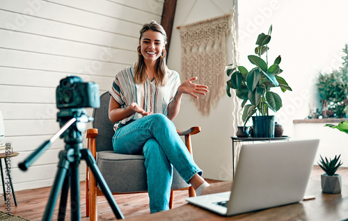 Attractive young woman blogger dressed in jeans and a blouse is recording on the camera while sitting on a comfortable chair at home. photo