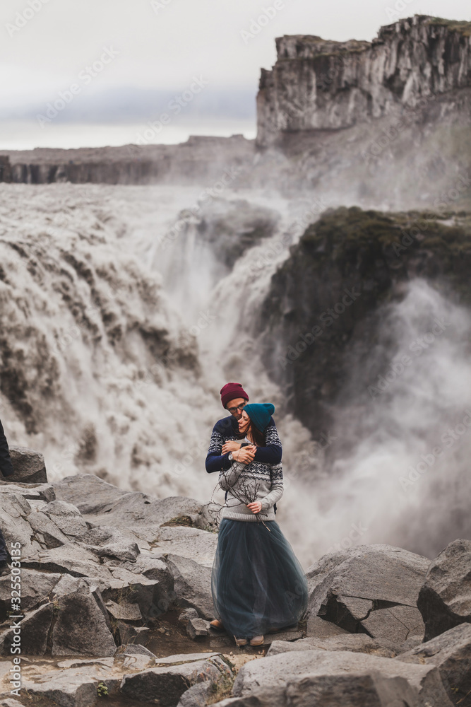 Close up lovely portrait of young hipster couple kissing together near famous Icelandic landscape - Dettifoss powerful waterfall. Traditional handmade wool sweaters, blue and red hats. Close up lovely portrait of young hipster couple kissing together near famous Icelandic landscape - Dettifoss powerful waterfall. Traditional handmade wool sweaters, blue and red hats.