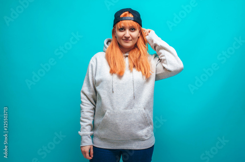 Handsome hipster female with bright red hair wears oversized gray blank hoodie and black cap with space for your logo or design on blue studio background.