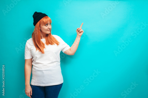 Hipster female with bright red hair wears white oversized t shirt pointing fingers at your text  isolated on blue studio wall. Crazy emotions. Discount  sale  season sales.