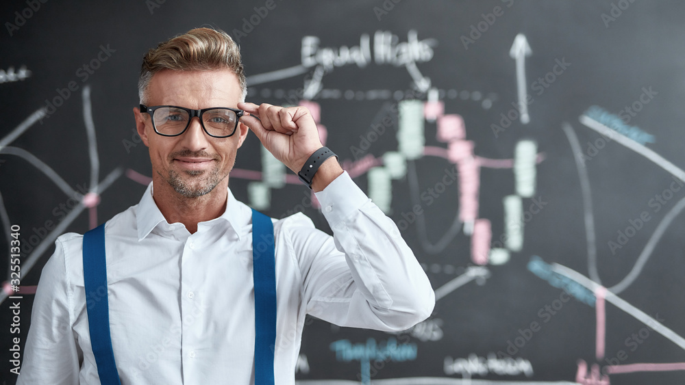 Broking for the source of income of tomorrow. Close up of middle-aged trader looking at camera and standing near blackboard full of charts, while thinking of the new strategy of work. Horizontal shot Broking for the source of income of tomorrow. Close up of middle-aged trader looking at camera and standing near blackboard full of charts, while thinking of the new strategy of work. Horizontal shot