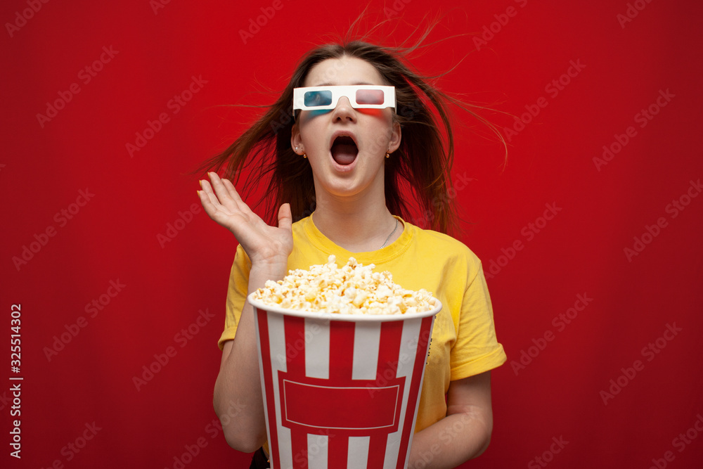 funny young girl student watching a movie in 3D glasses and screaming on a red background, she eats popcorn and is delighted